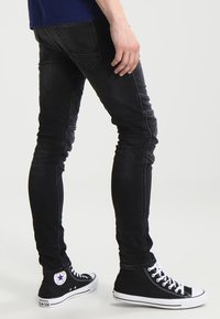 Mustat slim-fit farkut, joissa on ommeltuja yksityiskohtia ja kapenevat lahkeet, yhdistettynä mustiin korkeisiin tennareihin, joissa on valkoinen pohja ja logo.