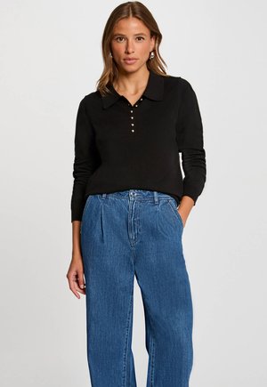 Zwarte coltrui met drie gouden knopen bij de halslijn, gecombineerd met hooggetailleerde blauwe denim wijde jeans. Gladde textuur.