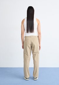 Beige cargo-bukser med sidelommer, afslappet pasform og lynlåsdetalje bagpå. Sammen med en hvid tanktop og sorte sneakers.