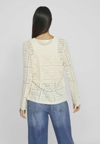 Suéter de punto en color crema con patrón de rombos abiertos, mangas largas, cuello redondo, corte relajado, combinado con unos vaqueros de pierna ancha azules.