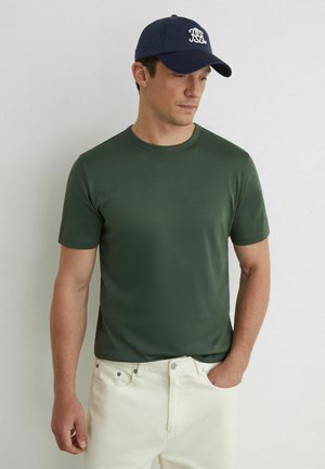 Mężczyzna w ciemnozielonej koszulce z krótkim rękawem, kremowych spodniach z ręką w kieszeni oraz granatowej czapce z daszkiem z białym logo, patrzący w dół w prawo.