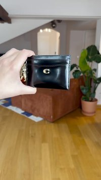Black leather cardholder with a glossy finish, featuring a gold "C" logo and multiple card slots, held against a wooden floor background.
