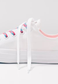 Zapatilla de lona blanca con superficie texturizada, con cordones rosa y azules, ojales redondos y suela de goma blanca.