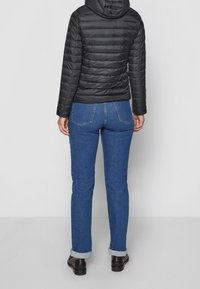 Person som bär en svart quiltad jacka med huva, blå manchesterjeans och svarta skor, stående mot en enkel ljus bakgrund.