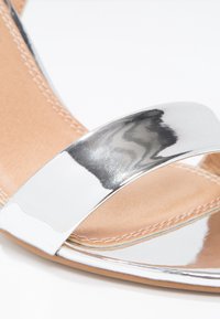 Sandale à talons métalliques argentés avec une large sangle sur le pied, révélant une semelle intérieure lisse couleur tan et des coutures détaillées le long du bord.