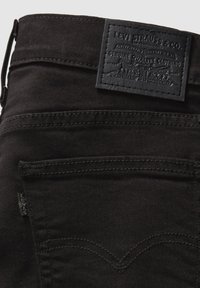 Jeans de mezclilla negra con una tela texturizada, que cuentan con un parche de cuero negro en la cintura y dos bolsillos traseros con detalles de costura.