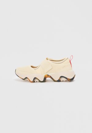 Lys beige glidelsko med teksturert overdel, justerbar stropp, chunky gummisåle med hvite og oransje detaljer, og en rød trekkløkke.