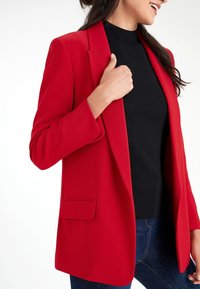 Rød skræddersyet blazer med hakkede revers og en enkelt forlomme, kombineret med en sort rullekrave og blå jeans. Glat materiale finish.