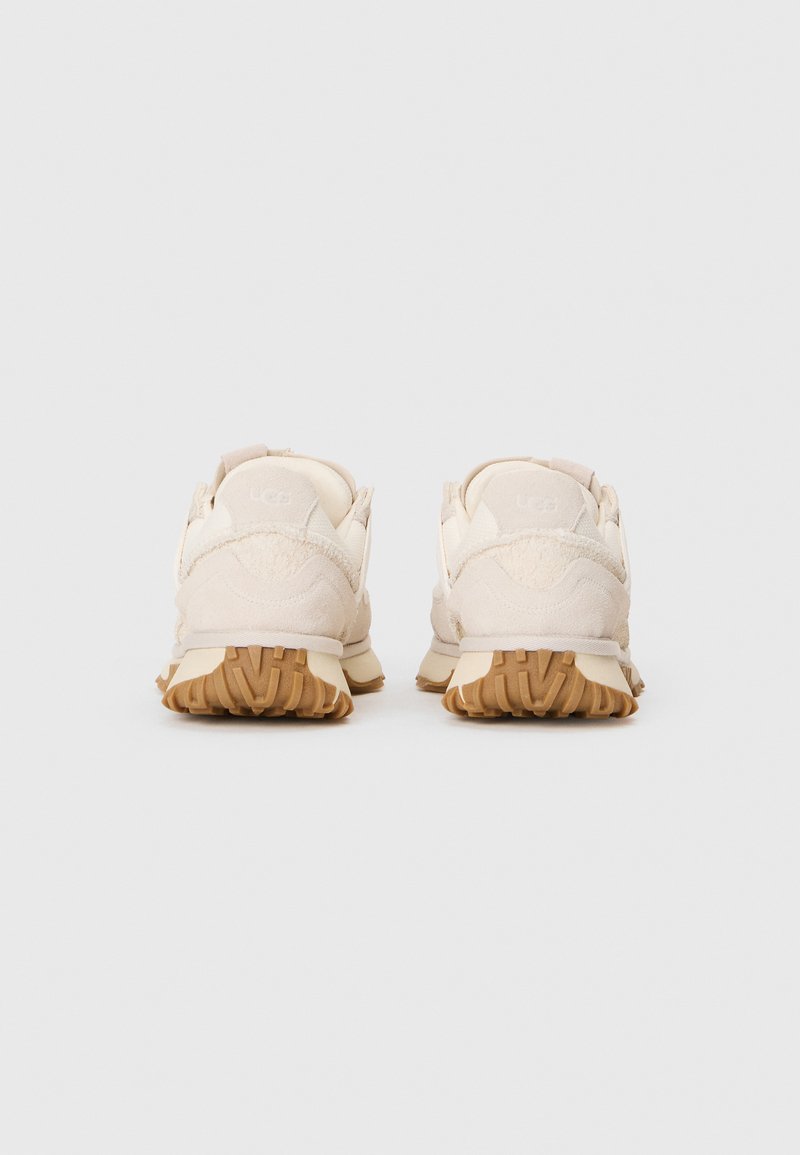 Beige Sneaker mit einer strukturierten Wildleder-Oberfläche, gepolstertem Absatz und Gummigummi-Sohle. Schlichtes Design mit minimalistischem Branding auf der Rückseite.