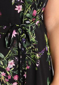 Musta mekko kukkakuviolla, jossa on vaaleanpunaisia ja purppuranvärisiä kukkia. Mukana on vyötäröllä solmittava nauha säädettävää istuvuutta varten. Silkkinen pinta.