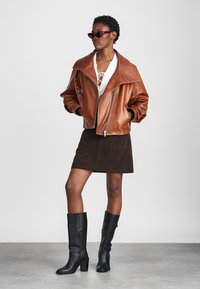 Brown leather jacket with wide collar, white lace-up top, brown suede mini skirt, and black knee-high boots. Dark sunglasses accent the look.