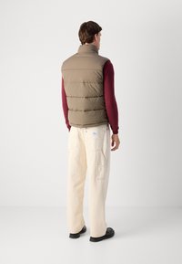 Man standing facing away, wearing a beige quilted vest, burgundy long-sleeve shirt, cream pants with pockets, and black shoes on a plain background.