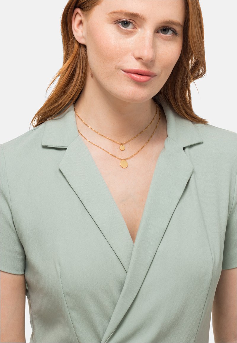 Abito avvolgente verde chiaro con maniche corte, caratterizzato da una superficie liscia e una profonda scollatura a V. Collana dorata a doppio strato con pendenti circolari.