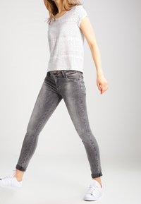 Grå stramme jeans med opslåede manchetter og en tekstureret hvid kortærmet top. Model iført hvide sneakers, poserer med det ene ben fremad.