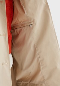 Intérieur d'un manteau beige avec une poche zippée et une partie d'un vêtement rouge et blanc visible en dessous.