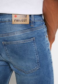 Modré džíny z denimu s koženou etiketou zobrazenou s nápisom "DENIM PROJECT." Majú štandardné zadné vrecká a viditeľné detaily šitia.