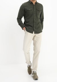 Olive green button-up shirt with two chest pockets, paired with light beige pants and brown sneakers with white accents.