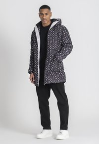 Chaqueta negra acolchada con estampado de logo blanco, cremallera y capucha. Lleva puesta sobre una camiseta negra con pantalones negros y zapatillas blancas.