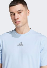 Světle modré sportovní tričko vyrobené z hladké, prodyšné tkaniny. Na hrudi se nachází stříbrné logo Adidas. Krátké rukávy, design s kulatým límcem.