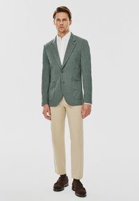 Jasnozielona kurtka z fakturowanej tkaniny, z klapami, dwoma przednimi kieszeniami, w połączeniu z beżowymi spodniami slim-fit i brązowymi butami. Pod spodem biała koszula.