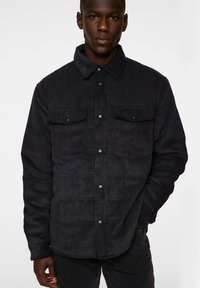 Svart kordflorskjorta med knäppning fram, två bröstfickor, ribbad textur och krage. Har en strukturerad passform.