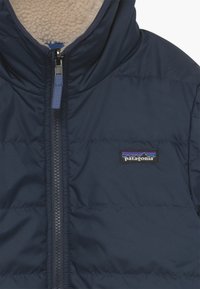 Marineblauwe gewatteerde jas met een hoge kraag, ritssluiting en fleece voering. Voorzien van een Patagonia-logo patch op de borst. Soepele textuur.