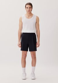 Top in cotone bianco con texture abbinato a pantaloni corti a pieghe blu navy. Il modello indossa calzini bianchi e sneakers, appoggiato a uno sfondo chiaro e neutro.