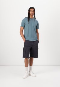 Camiseta de manga curta azul clara, calções cargo escuros com bolsos laterais, sapatilhas brancas e meias de cano curto. Conjunto simples e casual.