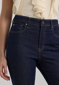 Tmavé džíny s vysokým pasem, se značkovým oranžovým šitím, zapínáním na knoflík a bočními kapsami. Texturovaná tkanina s hladkým povrchem.