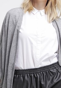 Cardigan grigio sopra una camicia bianca con bottoni, con colletto e leggero lucido. Shorts neri con cintura elastica e texture liscia.