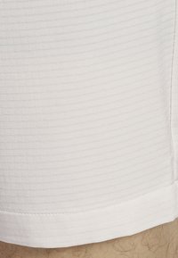 Gros plan sur l'ourlet de shorts blancs texturés au-dessus d'une jambe poilue, montrant le motif du tissu et les détails de couture.