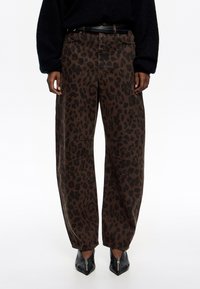 Hnedé nohavice so vzorom leopardích škvrn vyrobené z denimu, s vysokým pásom a zipsom. Čierne topánky s špicatou špičkou dopĺňajú vzhľad.