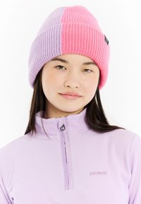 Gehaakte muts in roze en lavendel, met een geribbelde textuur en een kleurblokontwerp. Draagt een lichtpaarse fleecejas.