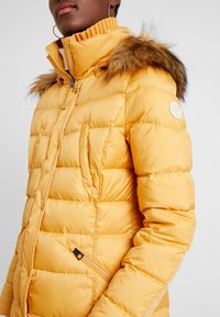 Woman wearing a mustard yellow quilted winter jacket with fur-trimmed hood and large hoop earrings, shown from chest to chin.