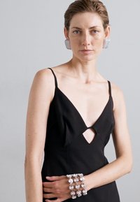 Schwarzes Kleid mit tiefem V-Ausschnitt und Frontausschnitt. Das Model trägt silberne Ohrringe und ein passendesperlenarmband mit runden Akzenten.
