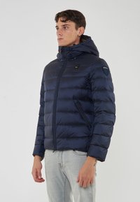 Giovane uomo che indossa una giacca a vento blu navy con cappuccio e tasche con zip, abbinata a jeans grigi chiari, rivolto verso sinistra.