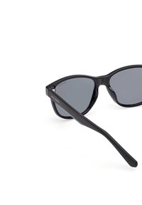 Svarta polariserade solglasögon med en spetsig kattögaform, slät plastbåge och gråtonade linser. Sidogången har "SKECHERS"-logotyp.