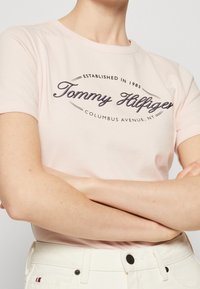Světle růžové tričko s kulatým výstřihem a krátkými rukávy. Má tmavý grafický logo s nápisem "Tommy Hilfiger" a rokem založení.