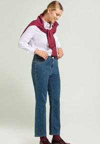 Blauwe denim jeans met een rechte pijp, hoge taille en vijf-pocket ontwerp, gecombineerd met een wit shirt en een bordeauxrode trui om de nek geknoopt.