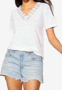 Žena ve bílém tričku s výstřihem do V a krajkovým lemem, v kombinaci s světle modrými roztrhanými džíny, zobrazená od ramen po polovinu stehna.