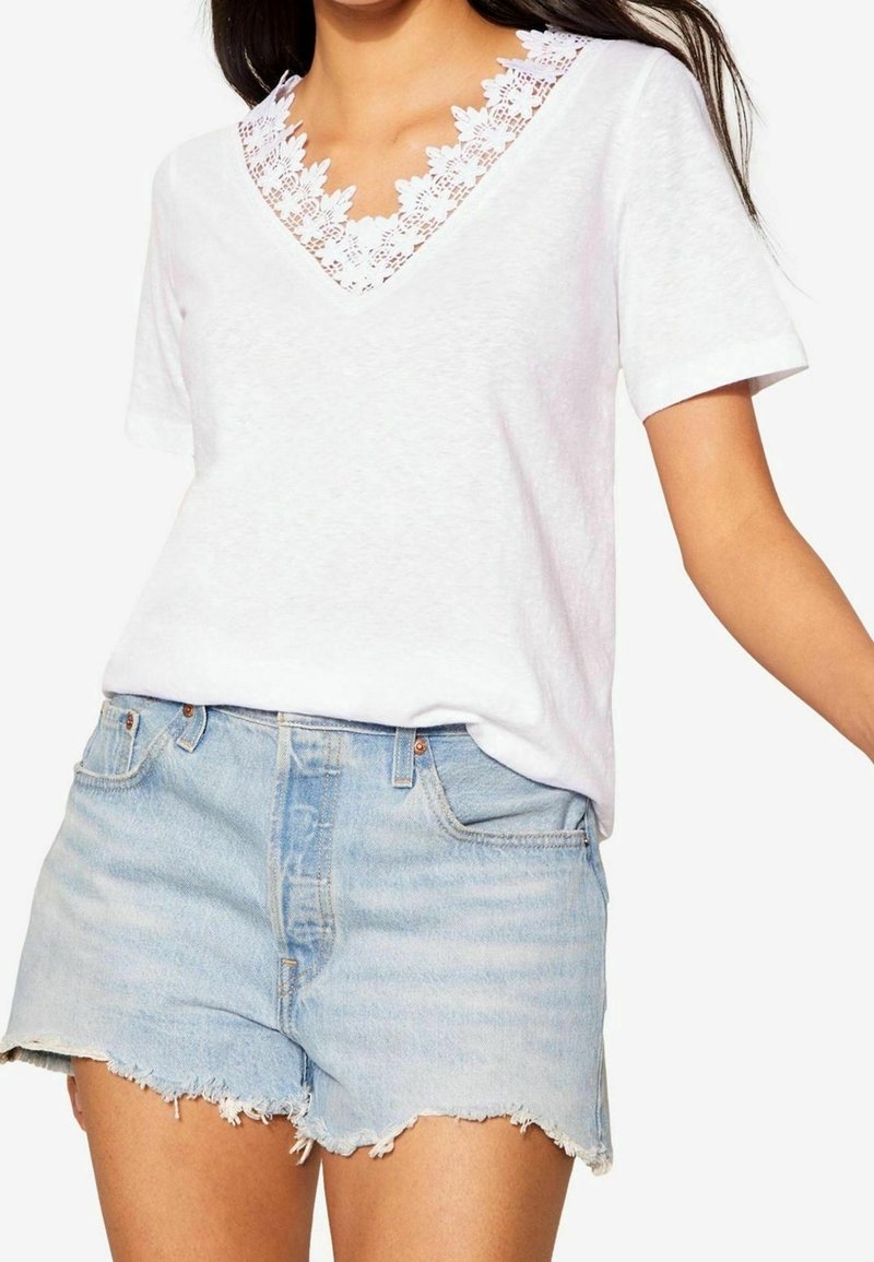 Žena ve bílém tričku s výstřihem do V a krajkovým lemem, v kombinaci s světle modrými roztrhanými džíny, zobrazená od ramen po polovinu stehna.