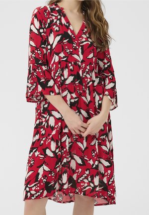 Frau trägt ein rotes knielanges Kleid mit schwarz-weißen Blumenmustern und dreiviertel Ärmeln, steht mit vor dem Körper verschränkten Händen.