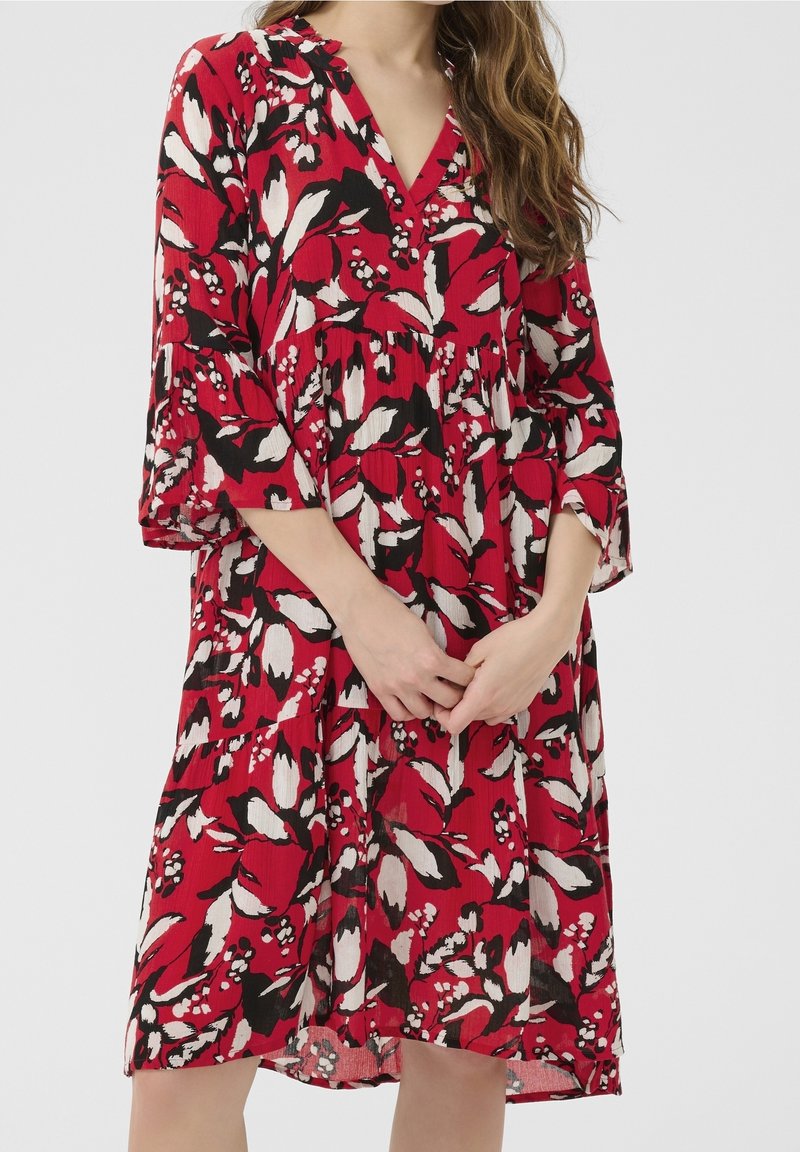 Femme portant une robe rouge au genou avec des motifs floraux noirs et blancs et des manches trois-quarts, debout, les mains jointes devant elle.