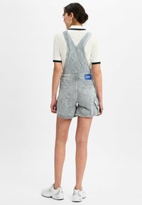 Vrouw staat met haar gezicht van de camera af, draagt gestreepte denim korte overall met een Hugo-label, een witte polo en witte sneakers.