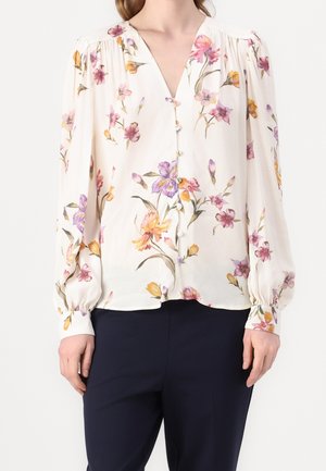 Hvid bluse med lilla, pink og gul blomsterprint, lange pufærmer, knaplukning foran, båret med mørke bukser.