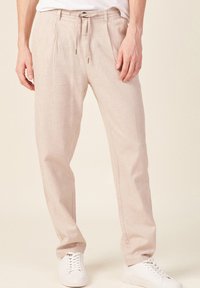 Pantalon en lin beige clair avec une coupe décontractée, doté d'une taille à cordon de serrage et de deux plis à l'avant. Porté avec des baskets blanches.