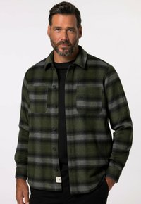 Camisa de franela a cuadros verde y gris con dos bolsillos en el pecho, cierre de botones y un dobladillo curvado. Camisa negra llevada debajo.