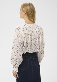 Blusa floral con mangas abullonadas, base color crema con pequeñas flores moradas, tela texturizada y detalle fruncido en la espalda, combinada con una falda de mezclilla oscura.