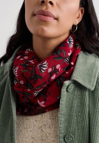 Sieviete, kura valkā zelta apli auskarus, sarkanu ziedu šalli, zaļu korduroja jaku un bēšu adītu džemperi, redzama no zoda līdz krūtīm.