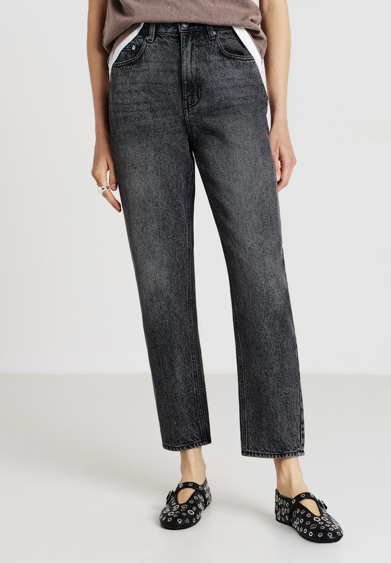 Persona che indossa jeans neri dritti lavati, scarpe Mary Jane nere con occhielli in metallo e una maglia marrone infilata all'interno su sfondo bianco.